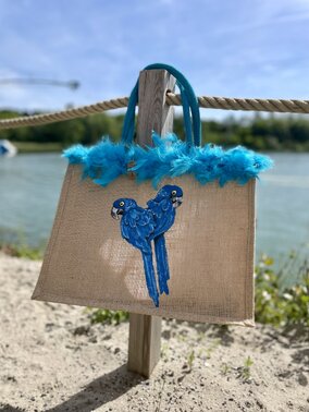 Sac de plage en jute avec des perroquets bleus peints à la main par Elles et No. et un boa en plumes collés sur le bord du sac